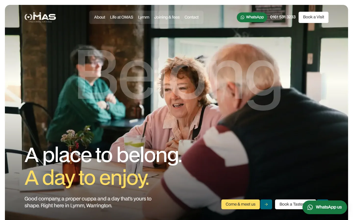 OMAS homepage: two older friends talking over drinks in a café, under the headline 'A place to belong. A day to enjoy.' with the word 'Belong' faint behind them